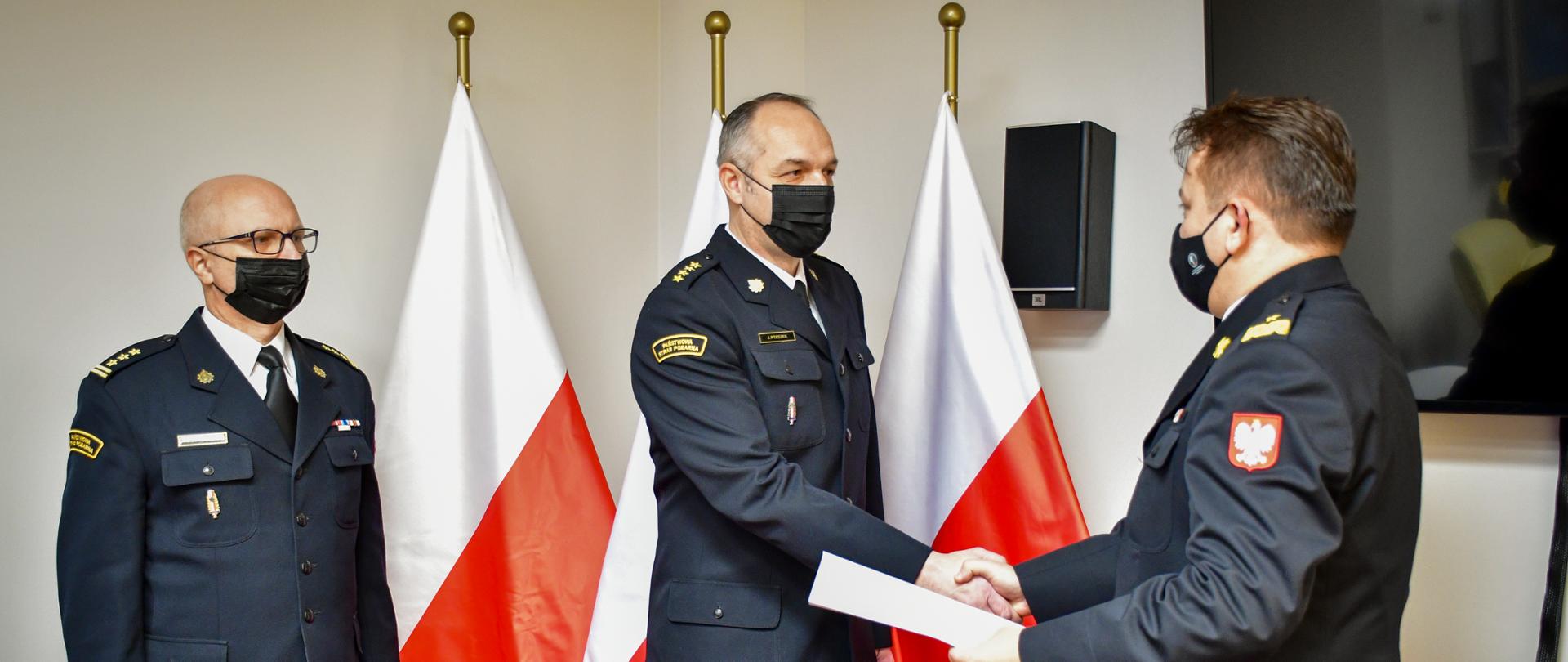 W pomieszczeniu stoi trzech mężczyzn ubranych w stroje wyjściowe PSP. Jeden mężczyzna podaje dłoń drugiemu trzymając w dłoni białą kartkę. Za mężczyznami są trzy flagi Polski.