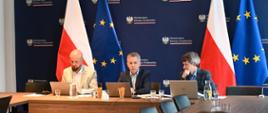 Trzy osoby siedzą z komputerami przy biurku. W centrum na tle flagi polskiej i EU siedzą członkowie Międzyresortowego Zespołu ds. Odry