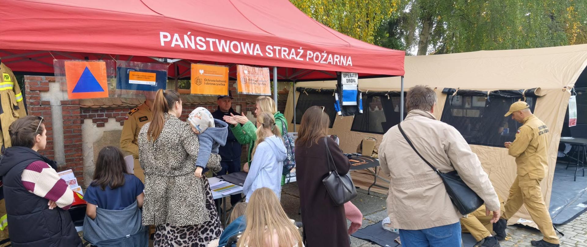 Zdjęcie przedstawia stoisko informacyjno-edukacyjne Państwowej Straży Pożarnej na świeżym powietrzu. Przed czerwonym namiotem strażackim oraz sąsiadującym z nim beżowym namiotem sztabowym zgromadziła się grupa dorosłych i dzieci, którzy rozmawiają z mundurowymi. Po prawej stronie ratownik w piaskowym mundurze demonstruje lub przygotowuje stanowisko do ćwiczeń na macie rozłożonej na ziemi. Całość odbywa się na wybrukowanym placu na tle jesiennych drzew.