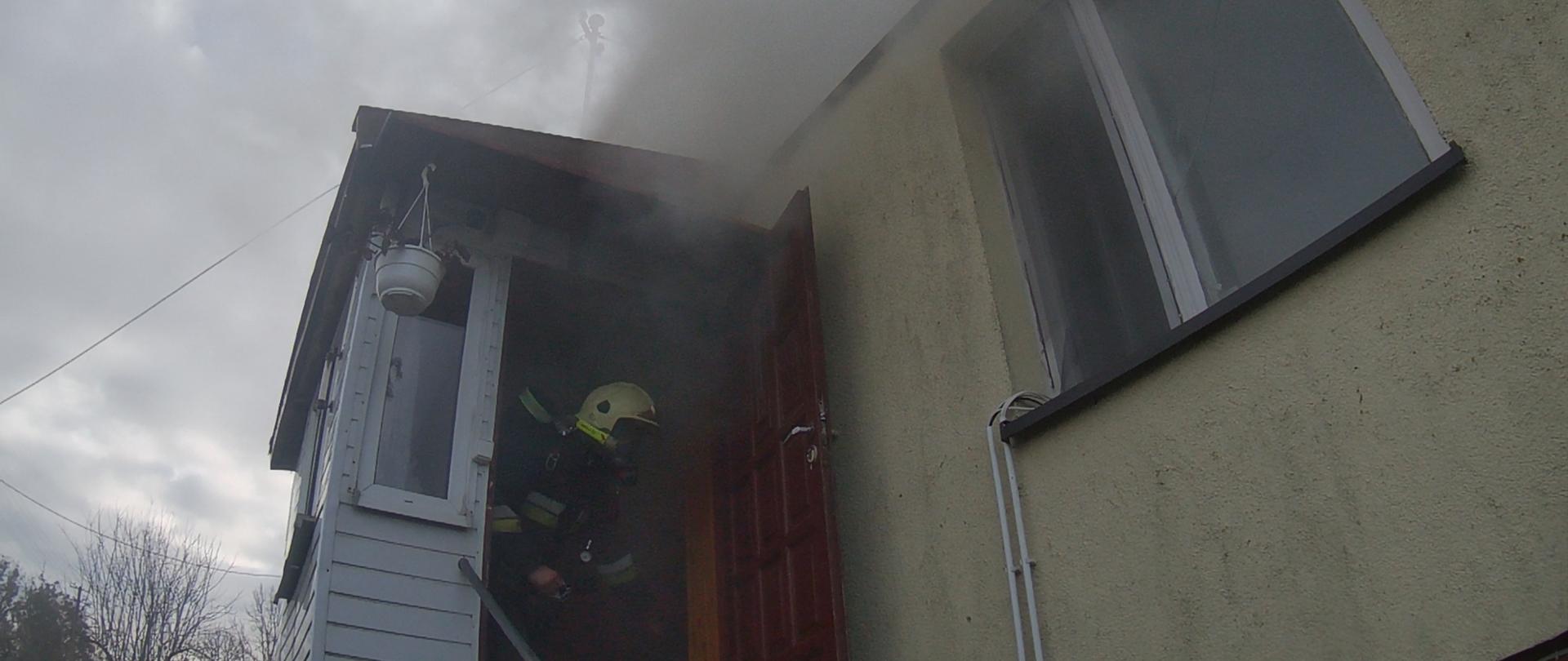 dom z którego wnętrza wydobywa się gęsty dym, w drzwiach wejściowych widać strażaka w aparacie ochrony układu oddechowego. Dzień.