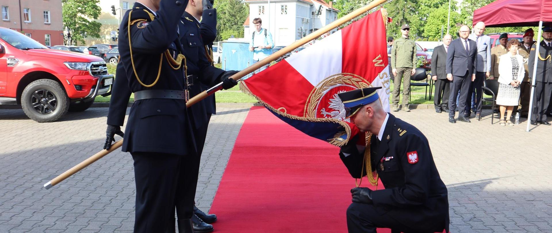 Po lewej stronie poczet sztandarowy. Sztandar Komendy Miejskiej Państwowej Straży Pożarnej w Toruniu skierowany w prawa stronę. Na środku zdjęcia rozwinięty czerwony dywan, na którym przyjmujący obowiązki Komendanta bryg. Sławomir Grudziński wita się ze sztandarem.