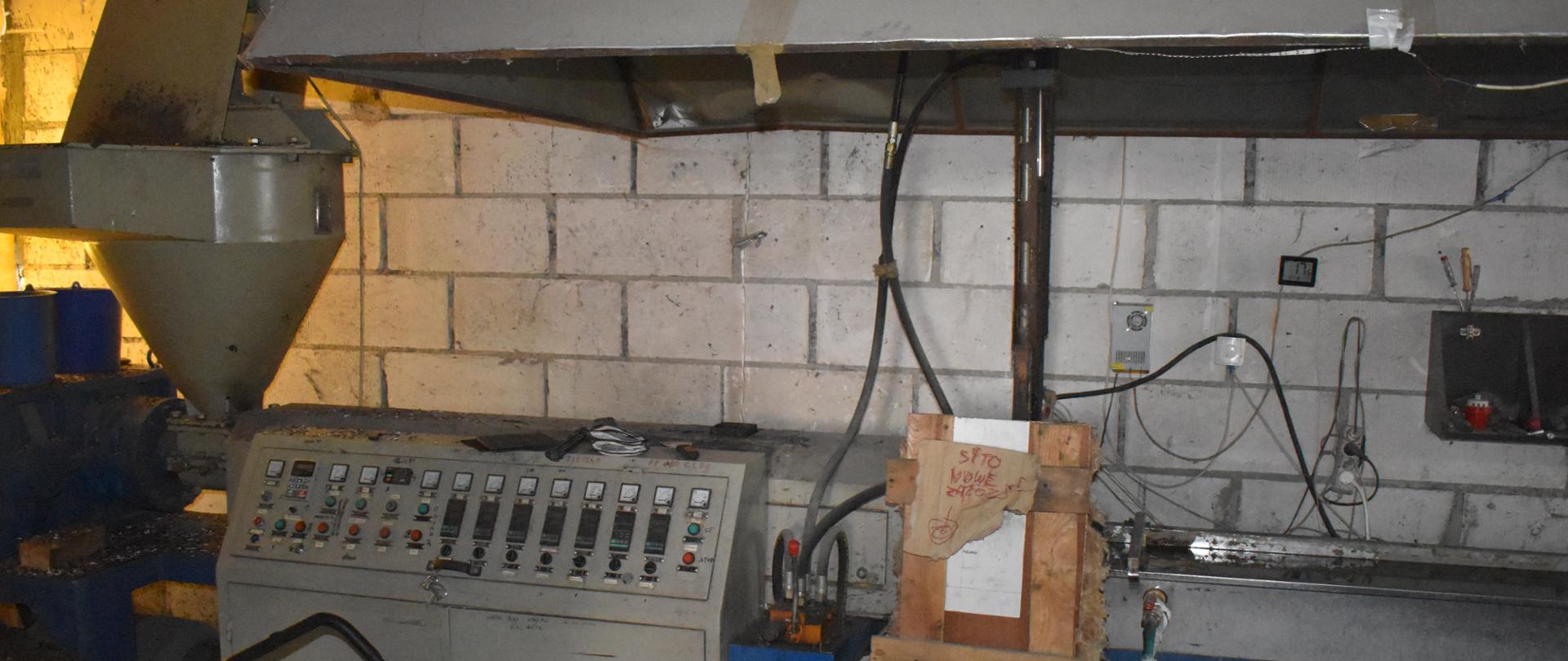 Stojąca w pomieszczeniu instalacja do przetwarzania odpadów wysoka na około 1 metr: po prawej stronie metalowy blat roboczy, po jego lewej stronie stalowa szafka z przełącznikami. Ponad instalacją znajduje się stalowy okap. Wszystkie urządzenia są w szarym kolorze. Bezpośrednio za nimi widać ścianę z białych cegieł.