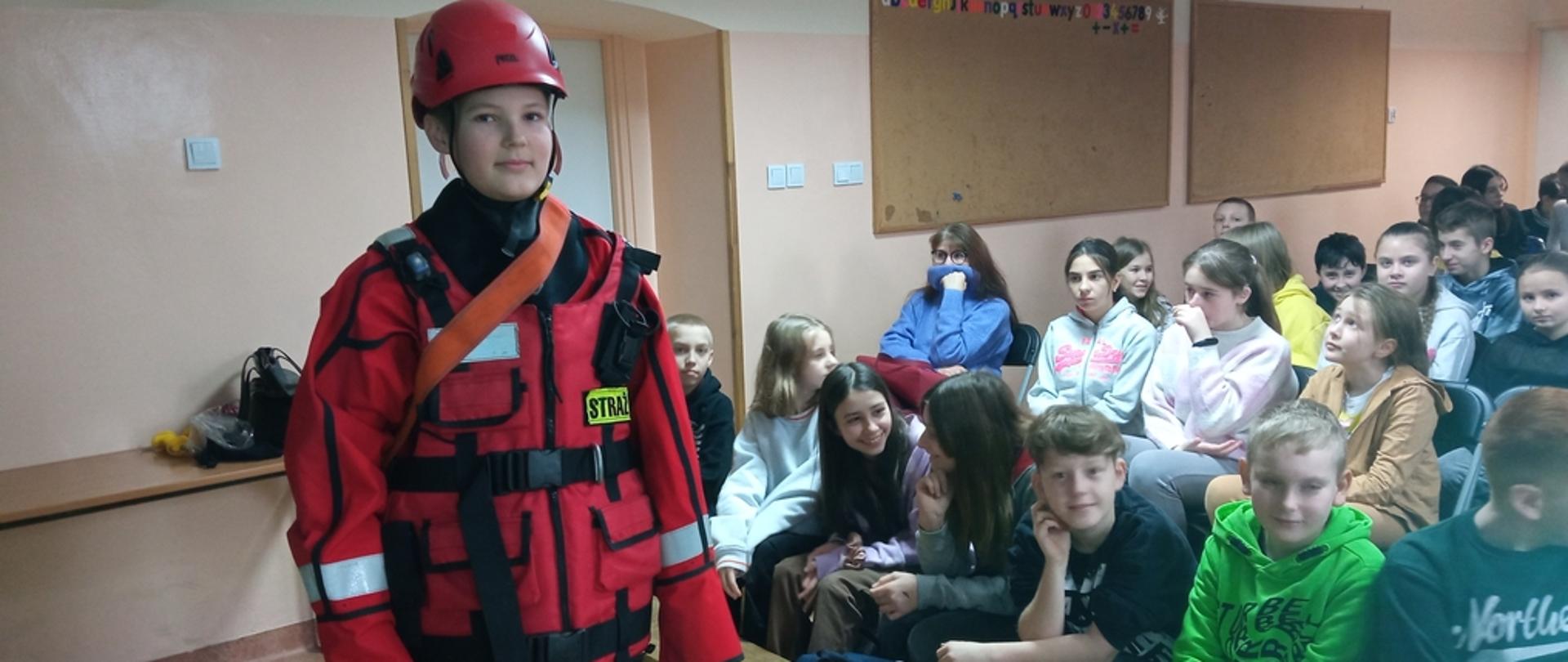 Na zdjęciu widoczne siedzące na krzesełkach dzieci. Jeden z chłopców ubrany w suchy skafander do działań w wodzie. Dzieci poznają sprzęt ratowniczy podczas prowadzonych przez strażaków zajęć z zakresu bezpieczeństwa nad wodą w ramach programu "Aktywni Błękitni - szkoła przyjazna wodzie"