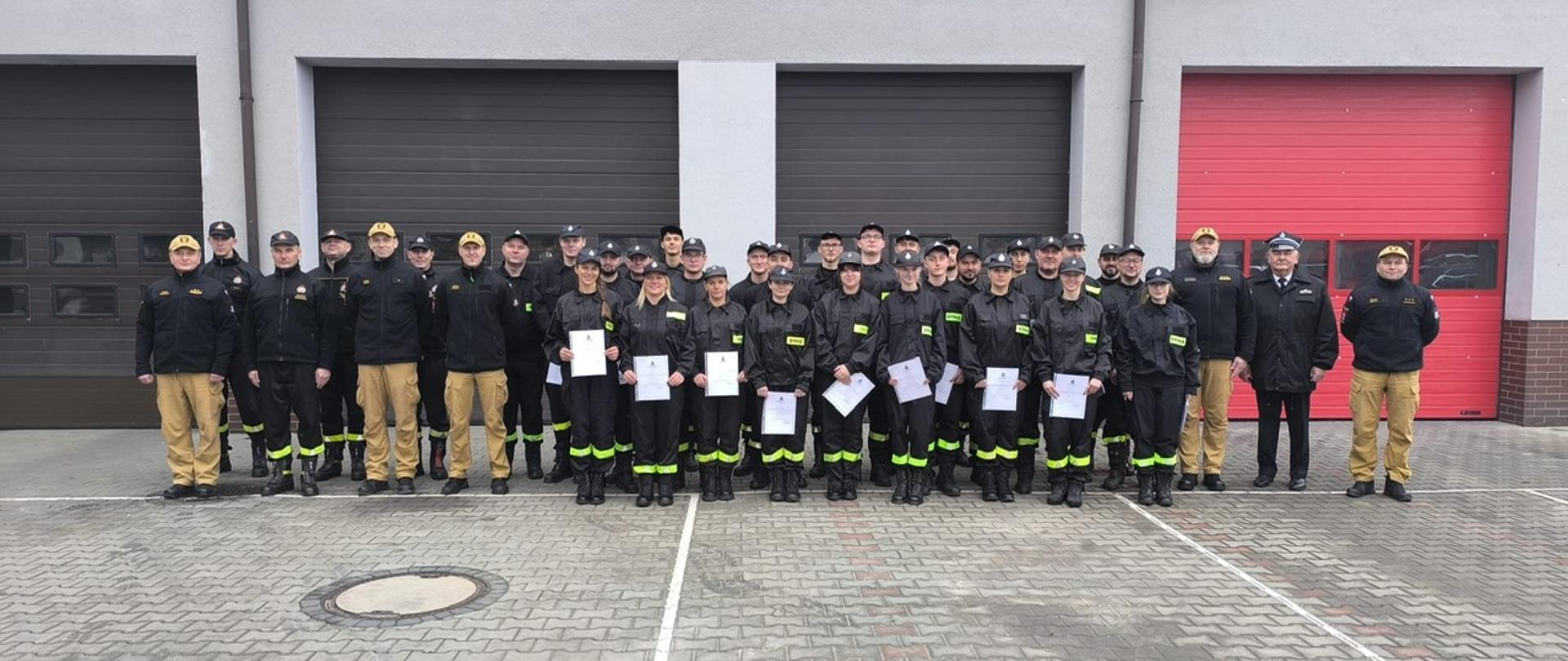 Zdjęcie przedstawia dużą grupę około 30-35 osób stojących rzędem przed budynkiem straży pożarnej. Większość osób ubrana jest w ciemne mundury strażackie (czarne kurtki i spodnie w kolorze khaki/beżowym), a część ma czarne czapki. W środkowej części rzędu stoi kilka kobiet i mężczyzn, którzy wydają się trzymać w rękach dokumenty lub certyfikaty, co sugeruje ukończenie szkolenia lub inną uroczystość.
Budynek za nimi ma szarą dolną część i różową/jasnoczerwoną górną. Widoczne są duże, zamknięte bramy garażowe (dwie szare po lewej i jedna czerwona po prawej). Na szarej ścianie umieszczone są klimatyzatory. Grupa stoi na wybrukowanym placu.