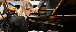 Zdjęcie przedstawiające pianistkę - Maję Klinowską na scenie filharmonii. W tle orkiestra symfoniczna. 