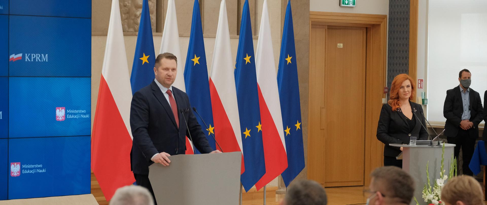 na śroku zdjęcia widać ministra Przemysława Czarnka przemawiającego do mikrofonu przy mównicy. Minister stoi na tle flag Polski i UE. z prawej strony zdjęcia stoi Rzecznik Prasowy MEiN Anna Ostrowska. 