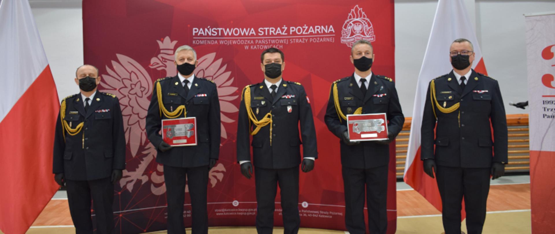 Śląski Komendant Wojewódzki Państwowej Straży Pożarnej nadbryg. Jacek Kleszczewski, zastępcy Komendanta Wojewódzkiego st. bryg. Bogdan Jędrocha, st. bryg. Mirosław Synowiec oraz funkcjonariusze odchodzący na zaopatrzenie emerytalne, stojący dla tle ścianki z godłem Polski i napisem Państwowa Straż Pożarna, po bokach dwie flagi państwowe. 