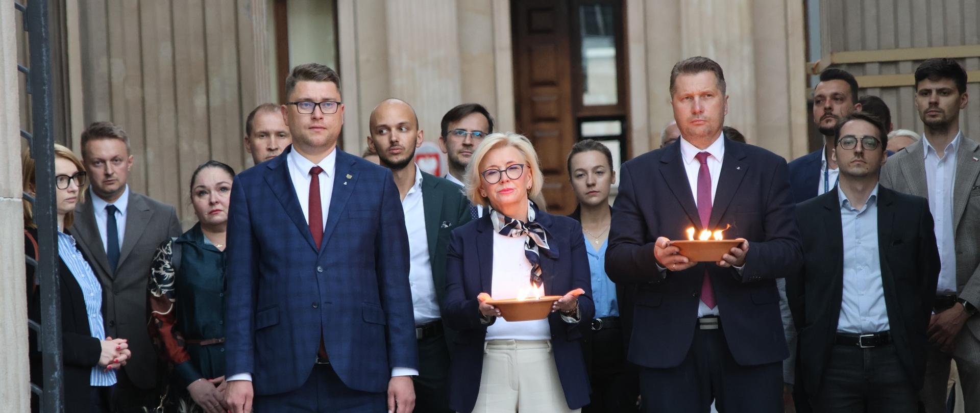 Przed wejściem do szarego budynku stoi grupa ludzi, z przodu minister Czarnek i wiceminister Machałek trzymają w rękach płonące znicze.