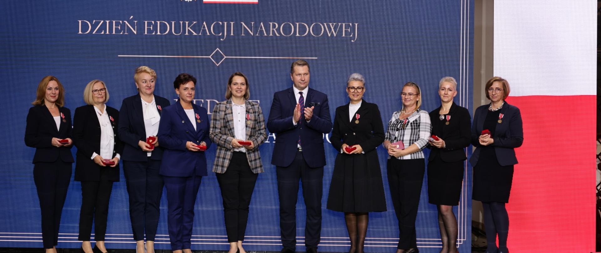 Pod niebieską ścianą z napisem Dzień Edukacji Narodowej stoi w szeregu grupa dziewięciu osób, trzymają małe czerwone pudełka.