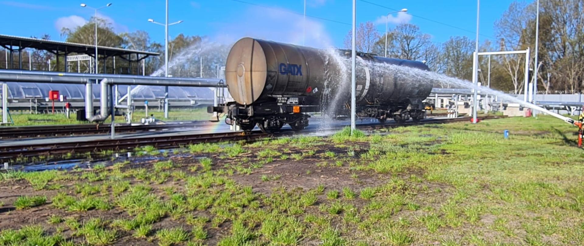 Zdjęcia przedstawia cysternę kolejową o ciemnym kolorze na torach oblewaną z dwóch stron działkami wodnymi. W tle widać instalację technologiczną. Na pierwszym planie zielona trawa.