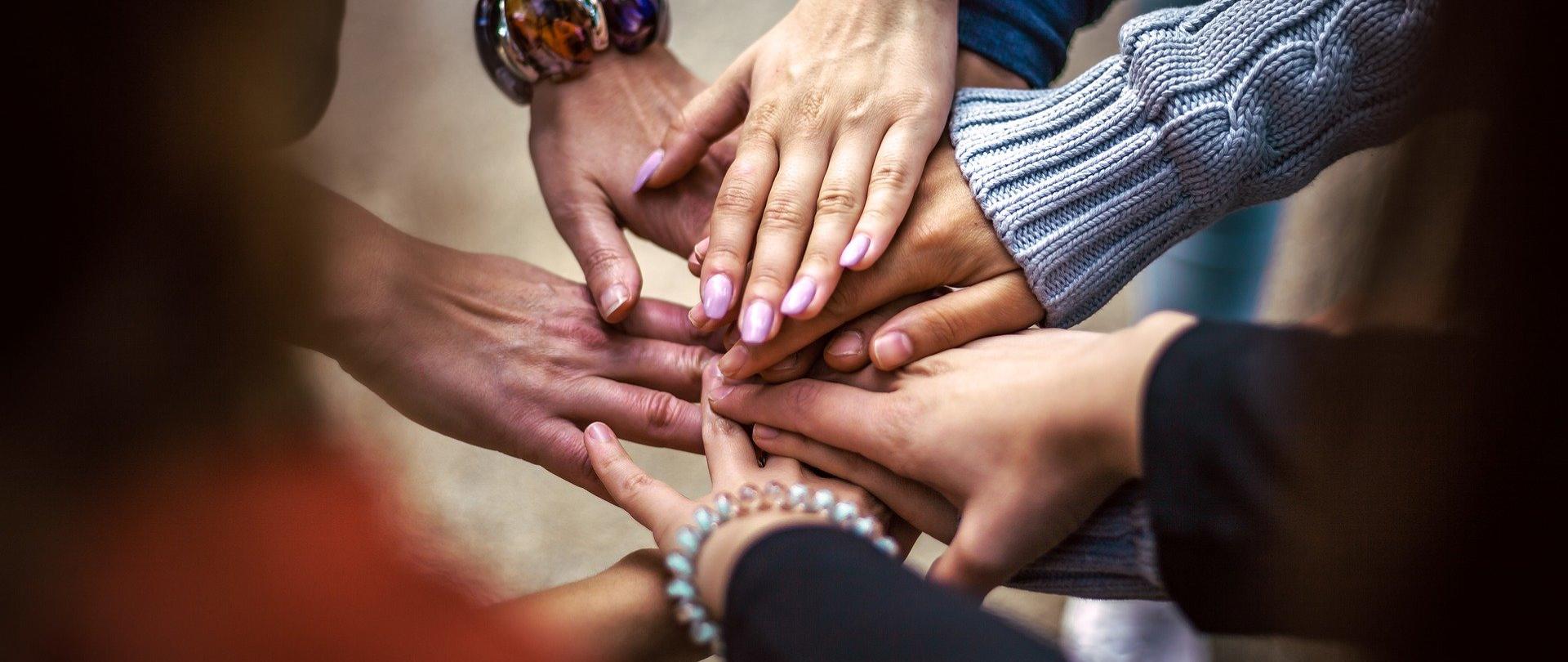 Zdjęcie przedstawia kilka par dłoni o różnym odcieniu skóry. Dłonie ułożone na znak współpracy - tj. jedna dłoń na drugą, w myśl zasady" jeden za wszystkich, wszyscy za jednego". 