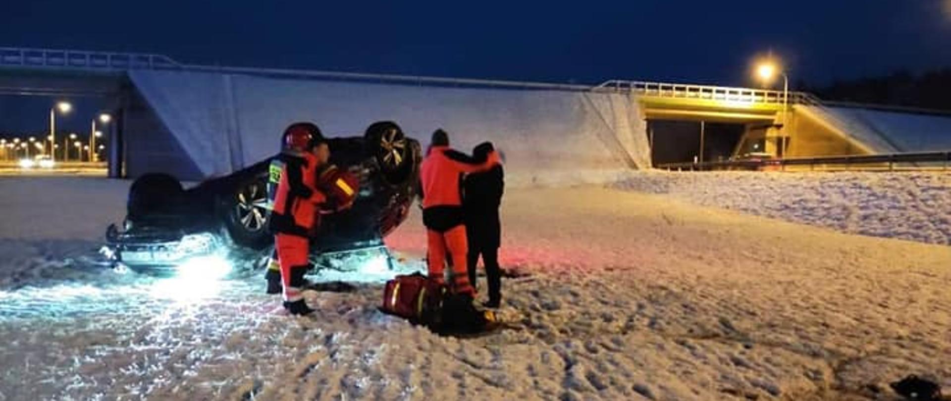 Zdjęcie przedstawia samochód na dachu oraz służby udzielające pomocy poszkodowanej. W tle wiadukt drogowy nad drogą ekspresową.