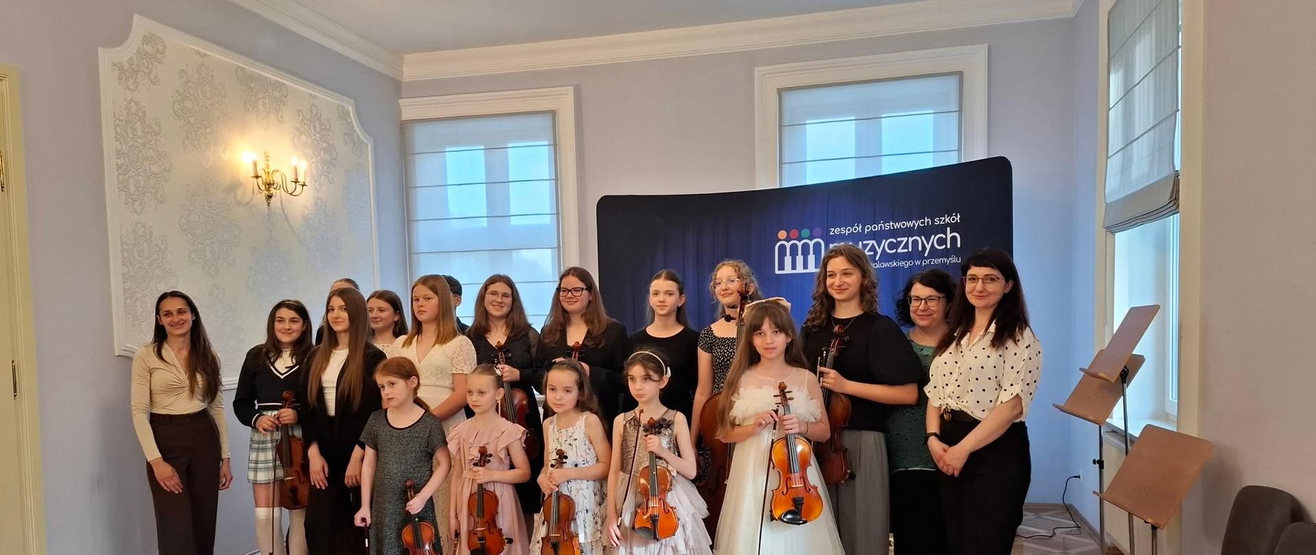 Zdjęcie przedstawia liczną grupę dziewcząt oraz dorosłych kobiet pozujących do wspólnej fotografii pamiątkowej w jasnej, klasycznej sali koncertowej z eleganckim drewnianym parkietem ułożonym w kasetony.
Na zdjęciu znajduje się kilkanaście młodszych i starszych uczennic klasy instrumentów smyczkowych, z których wiele trzyma w rękach swoje skrzypce oraz smyczki. Dziewczęta są ubrane w różnorodne, eleganckie stroje – od zwiewnych, tiulowych sukienek (w tym jedna dziewczynka w wyrazistej, białej kreacji w pierwszym rzędzie) po ciemne, galowe bluzki i spódnice u starszych uczennic. Po lewej stronie grupy stoi młoda kobieta w jasnej bluzce i ciemnych spodniach, natomiast po prawej stronie widać dwie dorosłe nauczycielki – jedna w ciemnej bluzce, a druga w białej koszuli w czarne grochy i czarnych spodniach.
W tle po lewej stronie znajduje się ozdobna ściana z klasycznym, jasnym motywem roślinnym i złotym kinkietem. W centralnej części, bezpośrednio za uczestniczkami, ustawiona jest łukowata, ciemnoniebieska ścianka z białym napisem „zespół państwowych szkół muzycznych im. artura malawskiego w przemyślu” oraz kolorowym logotypem. Po prawej stronie sali, w głębi, widoczne są wysokie okna, metalowe pulpity na nuty oraz rządem ustawione krzesła dla widowni.