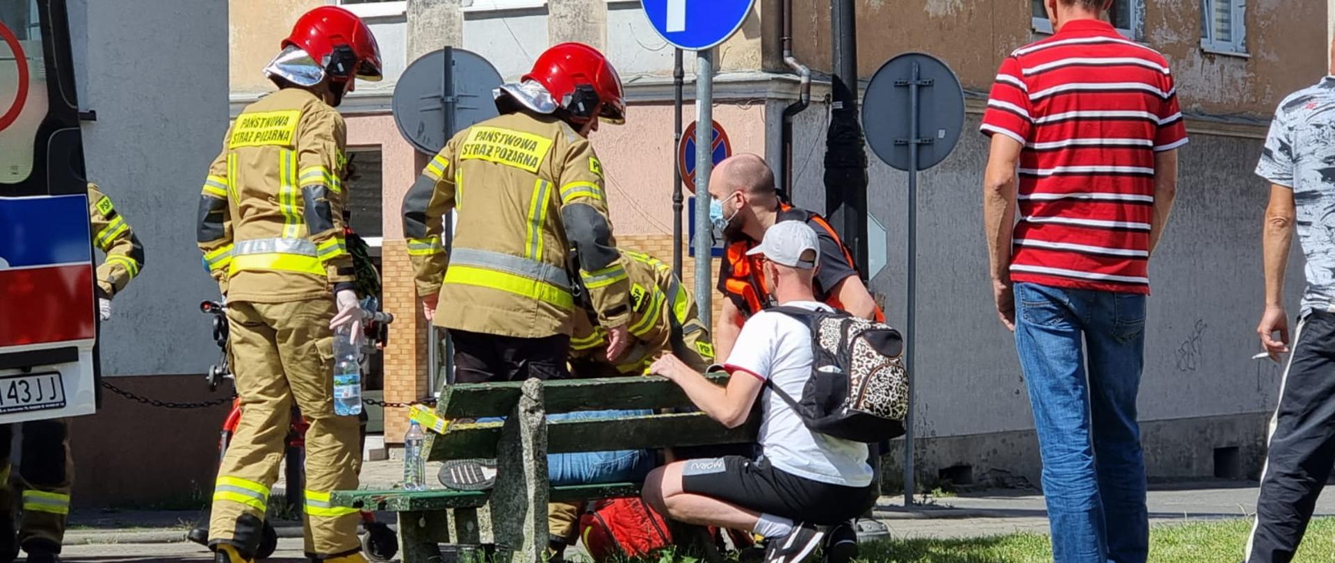Zdjęcie przedstawia strażaków udzielających pomocy osobie która zasłabła na ławce.Widać tez osoby postronne.