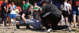Zdjęcie przedstawia młoda dziewczynę i młodego chłopaka udzielających pomoc osobie. Dziewczyna ma na dłoniach rękawiczki lateksowe, trzyma osobę poszkodowaną za nogi, chłopak trzyma poszkodowanego za tułów od strony głowy. W tle osoby przyglądające się akcji ratunkowej.