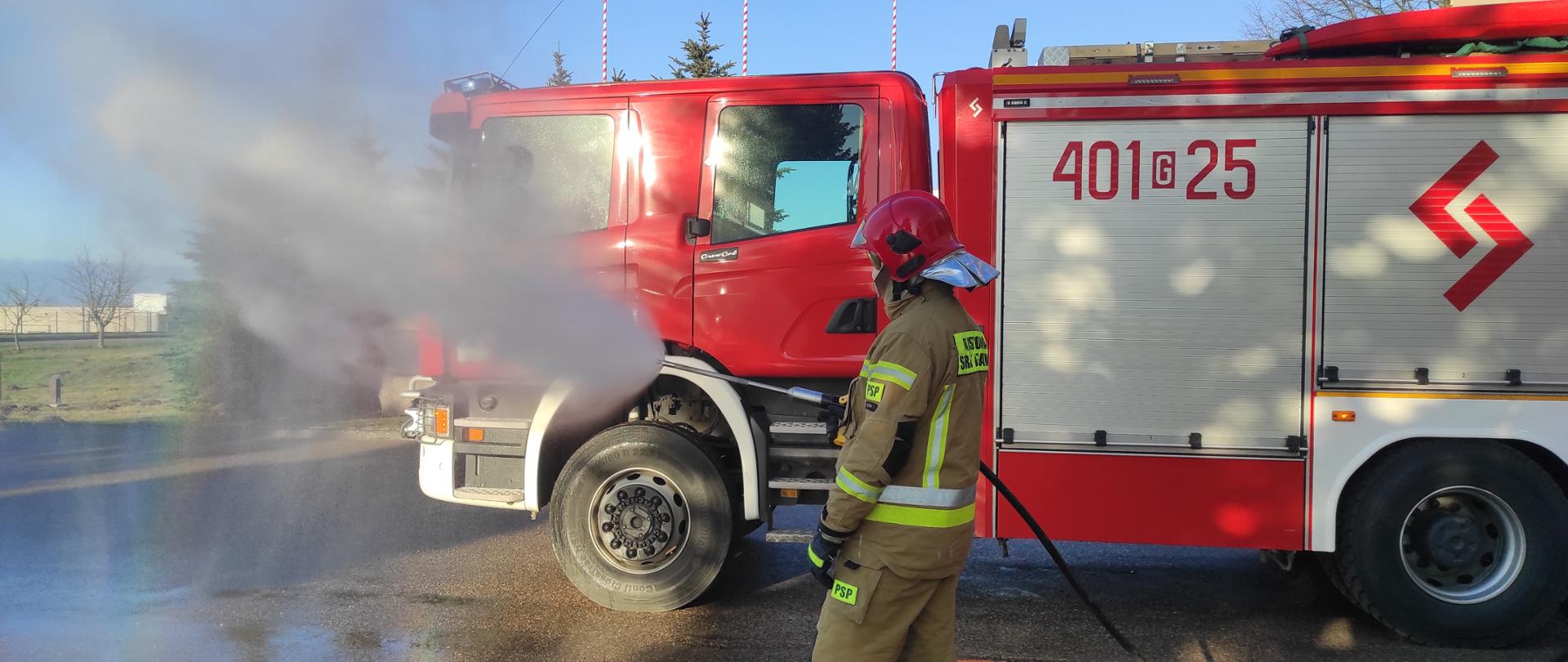 Na zdjęciu znajduje się strażak w czerwonym hełmie w umundurowaniu specjalnym koloru musztardowego, który leje wodę przy wykorzystaniu lancy gaśniczej. Za strazakiem ustawiony jest samochód ratowniczo-gaśniczy.