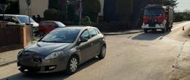 Po lewej stronie widać samochód fiat brawo, który brał udział w kolizji, po prawej samochód strażacki, w tle dom jednorodzinny, tuje, oraz stojące na poboczu samochody, 