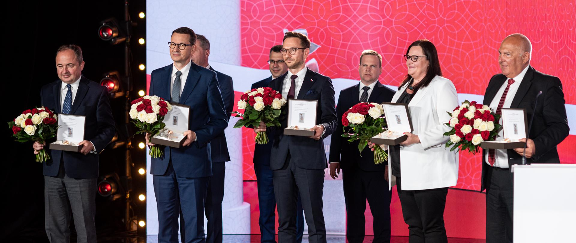 Na zdjęciu laureaci Biało-Czerwonych Róż portalu wPolityce.pl Premier Mateusz Morawiecki, minister ds. Unii Europejskiej w Kancelarii Prezesa Rady Ministrów Konrad Szymański, minister finansów, funduszy i polityki regionalnej Tadeusz Kościński, wiceminister funduszy i polityki regionalnej Małgorzata Jarosińska-Jedynak, wiceminister funduszy i polityki regionalnej Waldemar Buda stojący na scenie z nagrodami. 