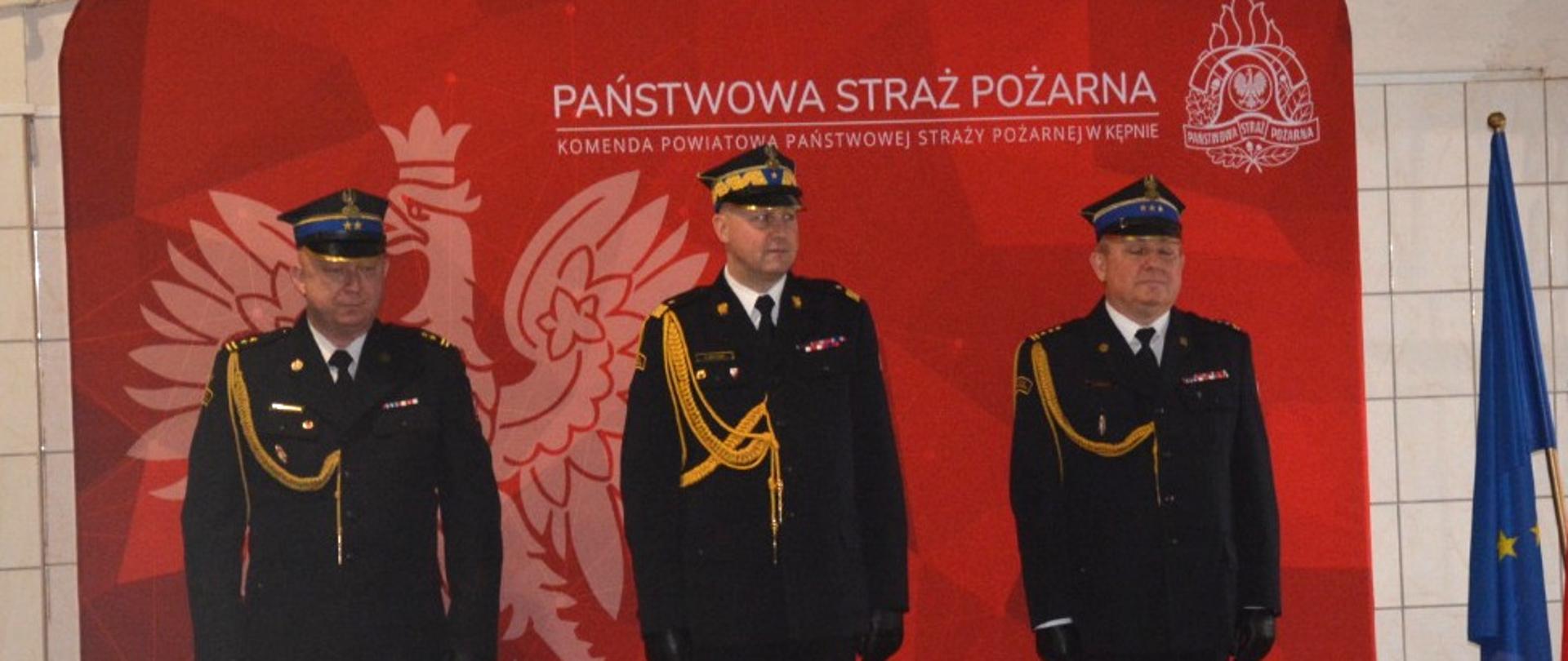 trzech strażaków stoi na baczność, za nimi czerwona ścianka z orłem i napiesem państwowa straż pożarne, po prawej stronie mównica przy której stoi kobieta 