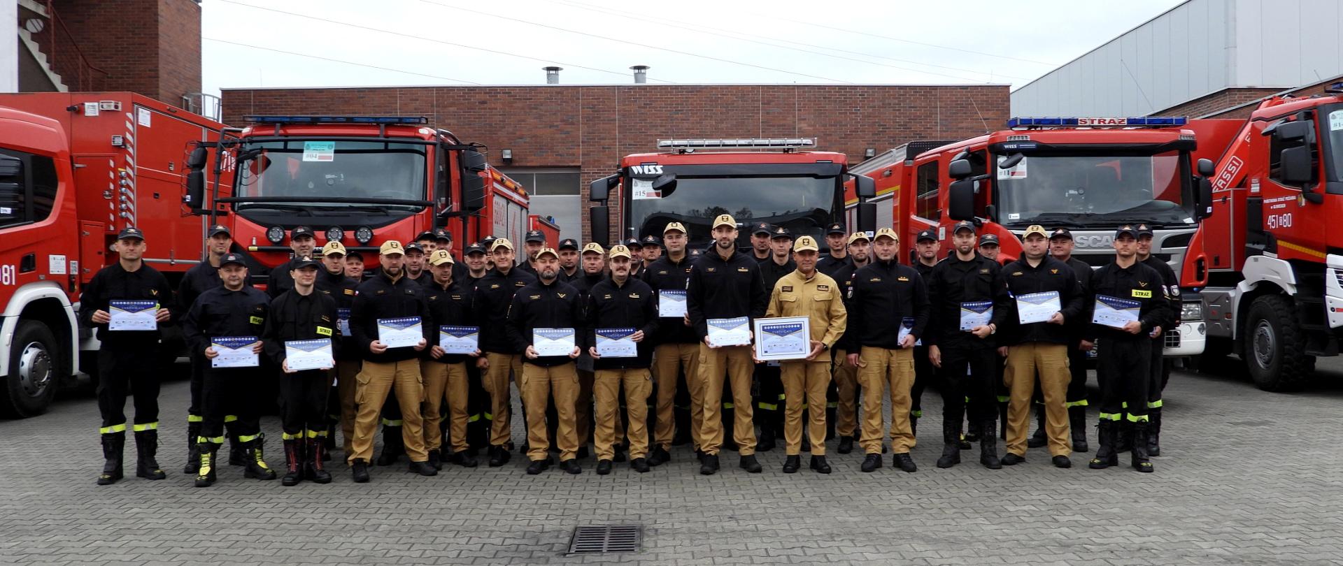 Na placu jednostki ratowniczo - gaśniczej w Katowicach - Szopienicach ustawiona do grupowego zdjęcia, grupa strażaków, w służbowych mundurach, z polskiego modułu pomp wysokiej wydajności oraz zastępca śląskiego komendanta wojewódzkiego PSP bryg. Arkadiusz Krzemiński. W rękach trzymają certyfikaty udziału w ćwiczeniach Modex. W tle, ustawione przodem samochody pożarnicze i ceglany budynek jednostki.