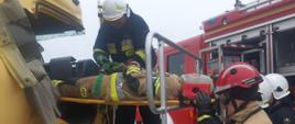 Warsztaty z zakresu ratownictwa technicznego. Złomowisko w Chojnie. Strażacy ewakuują z kabiny pojazdu na noszach typu deska poszkodowanego mężczyznę. Jeden z ratowników znajduje się na podeście ratowniczym - zabezpiecza ratowanego pasami.