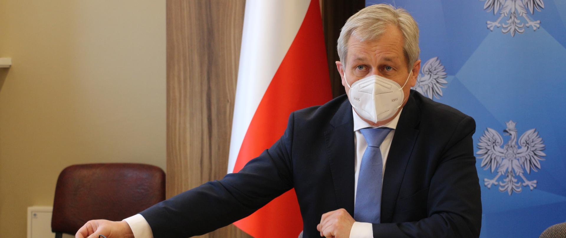 Na zdj. siedzący za stołem wojewoda opolski, oparty jedną ręką o stół. W dłoni trzyma okulary. Drugą dłoń ma zawiniętą w pięść, trzyma ją bliżej tułowia. Przed wojewodą na stole dokumenty. W tle roll up z napisem wojewoda opolski. 