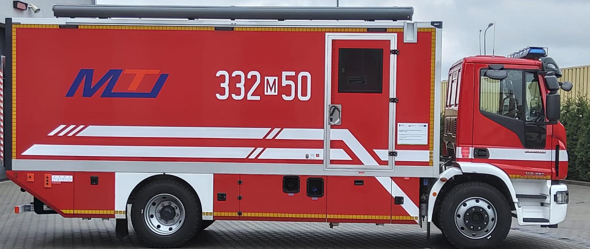 Zdjęcie przedstawia pojazd przed garażami jednostki ratowniczo – gaśniczej Państwowej Straży Pożarnej numer 2 w Radomiu. Na zdjęciu przedstawiono widok prawej strony samochodu. Pojazd jest w kolorze czerwonym ze srebrnymi i białymi elementami zabudowy. Za kabiną załogi znajduje się część warsztatowa przeznaczona do wykonywania bieżących prac serwisowych sprzętu ochrony dróg oddechowych oraz napełniania butli sprężonym powietrzem.