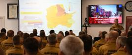 Strażak stoi przy mównicy i przemawia do funkcjonariuszy którzy siedzą na krzesłach na ekranach wyświetlana jest prezentacja.