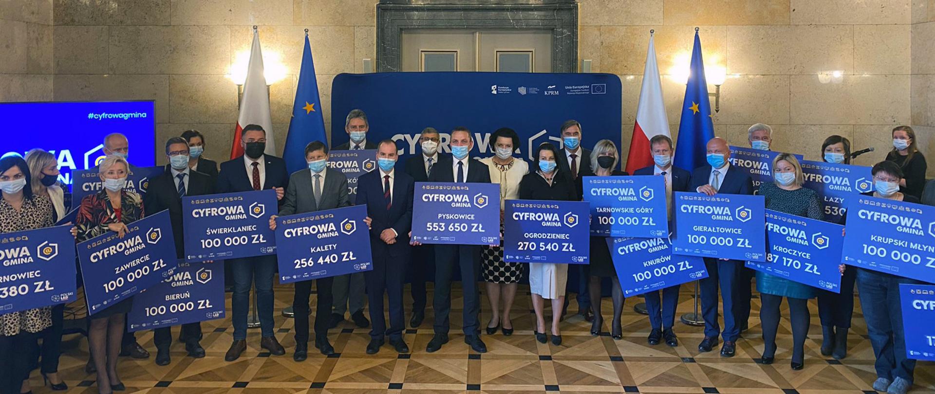 Minister Adam Andruszkiewicz na spotkaniu z samorządowcami ze Śląska w ramach projektu Cyfrowa Gmina. Samorządowcy trzymają symboliczne czeki. 