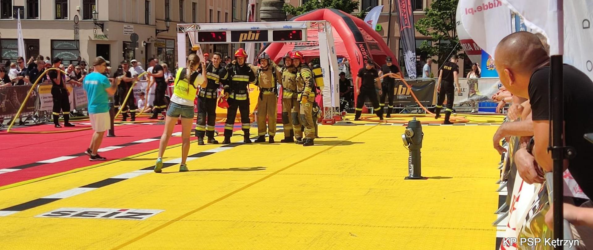 Pięciu strażaków ubranych w ubrania specjalne granatowe oraz piaskowe. Ustawieni do zdjęć na matach toru żółtego.