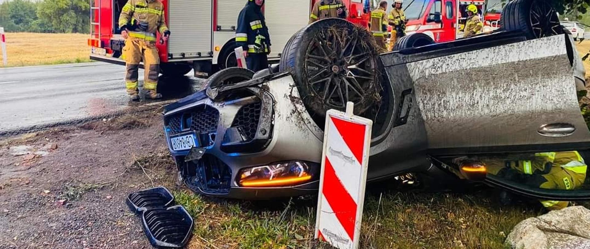 Pochmurno. Pobocze drogi. Srebrne auto leży na dachu , ma otwarte przednie drzwi przy których kuca strażak w jasnym mundurze. Obok na asfaltowej mokrej drodze stoją czerwone wozy strażackie. Przed nimi strażacy.