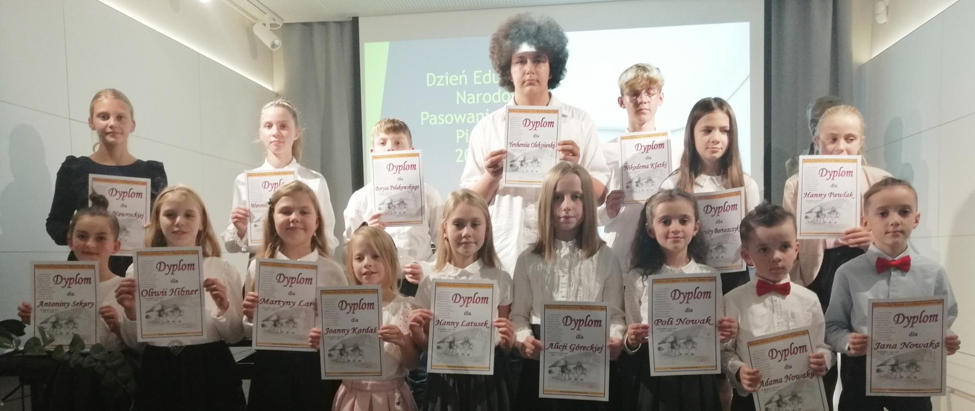 Zdjęcie przedstawia uczniów w strojach galowych stojących na stopniach sceny w dwuszeregu i trzymających w dłoniach dyplomy widoczne dla publiczności, upamiętniające Pasowanie na Ucznia. W tle na opuszczonym ekranie wyświetlona jest grafika w jasnych kolorach (biało-szaro-zielonym) z napisem "Dzień Edukacji Narodowej Pasowanie na Ucznia", który częściowo zasłaniają stojący uczniowie. 