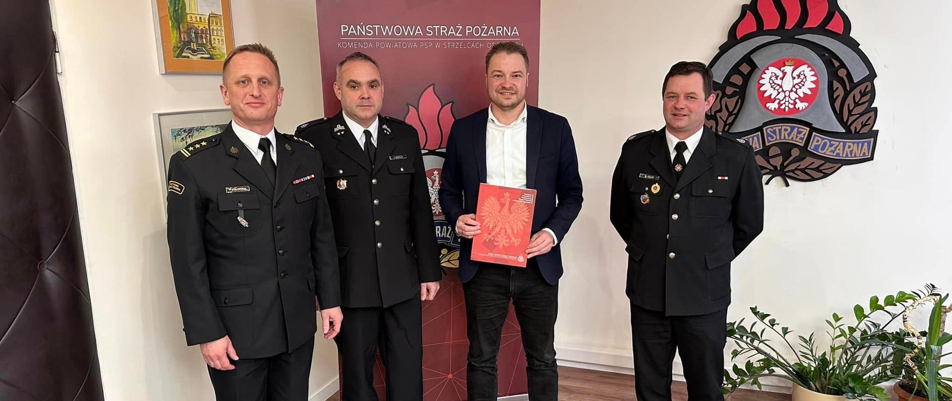 Na zdjęciu znajdują się trzej strażacy ubrani w mundury oraz osoba cywilna, w tle widać logo PSP na ścianie oraz baner