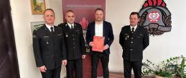 Na zdjęciu znajdują się trzej strażacy ubrani w mundury oraz osoba cywilna, w tle widać logo PSP na ścianie oraz baner