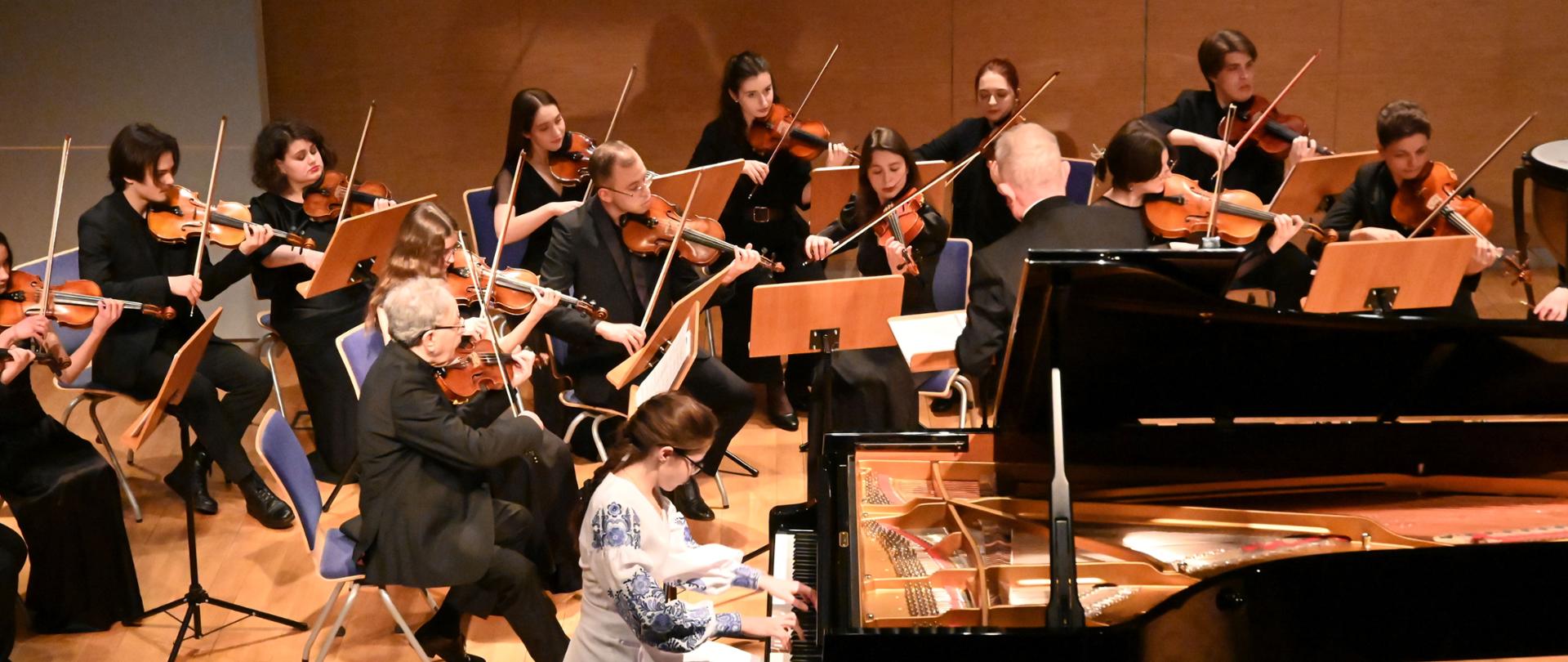 Widok z góry na orkiestrę smyczkową i pianistkę podczas występu. Młoda kobieta w białej sukni z niebieskim haftem (wzór ludowy) gra na otwartym czarnym fortepianie. Za nią siedzi kilkunastoosobowa orkiestra, a dyrygent stoi tyłem do widza, prowadząc zespół.