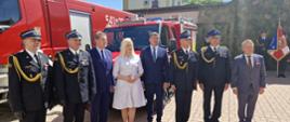 Zdjęcie przedstawia siedem osób stojących w rzędzie na brukowanym placu, w tle czerwony wóz strażacki i fragment budynku. Od lewej strony widocznych jest dwóch strażaków w galowych mundurach z dystynkcjami i medalami. Obok nich stoi mężczyzna w granatowym garniturze, a następnie kobieta w jasnoszarej sukience z kołnierzykiem. Kolejny jest mężczyzna w ciemnym garniturze, a za nim dwóch kolejnych strażaków w galowych mundurach. Ostatni w rzędzie to mężczyzna w szarym garniturze. Po prawej stronie zdjęcia, w tle, można dostrzec fragmenty kolejnych postaci w mundurach i sztandar. Niebo jest bezchmurne i błękitne.
