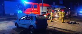 Widok na miejsce zdarzenia z drugiej strony. Centralny punkt zdjęcia stanowi rozbite auto osobowe koloru niebieskiego. W tle czerwony pojazd gaśniczy straży pożarnej i strażacy.