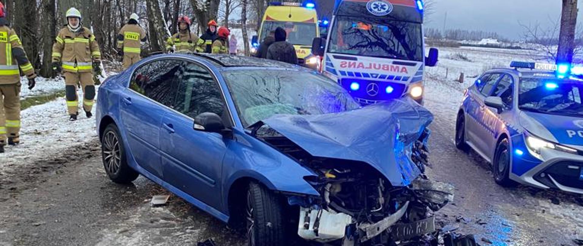 Na środku drogi stoi rozbity pojazd osobowy. niebieskie auto ma całkowicie zniszczony przód. Za nim widoczne sylwetki służb ratowniczych oraz pojazd pożarniczy, radiowóz oraz karetka pogotowia.