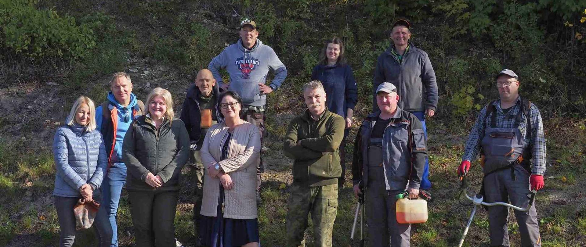 Grupa ludzi na tle zieleni. Ubrani są terenowo, niektórzy trzymają sprzęt do wykaszania