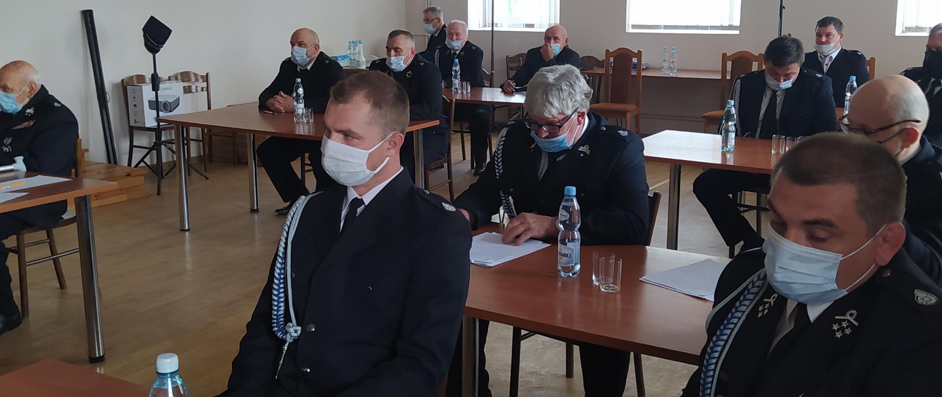 Druhowie OSP siedzą przy stolikach. Mają założone maseczki i. Ubrani są w mundury wyjściowe