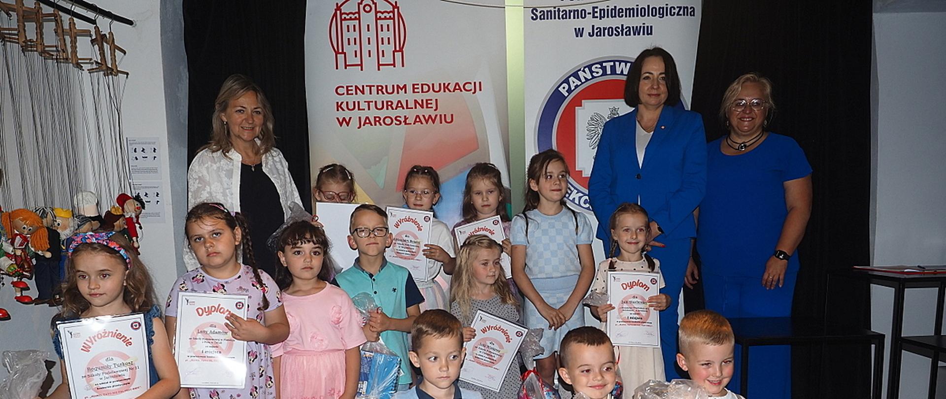 Grupa dzieci pokazuje swoje dyplomy, obok trzech kobiet na tle tła.