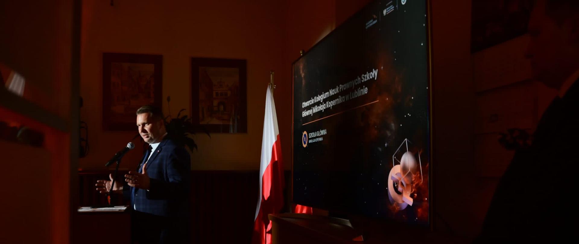 Zdjęcie z boku, w mrocznej sali za mównicą stoi minister Czarnek i mówi do mikrofonu.