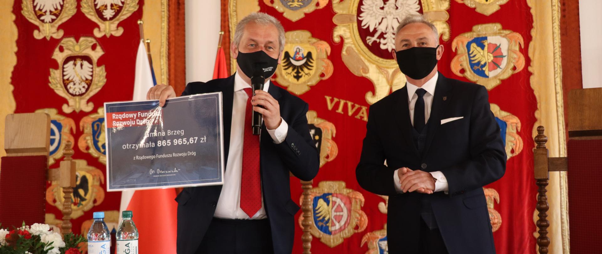 Na zdjęciu, stojący za stołem drewnianym wojewoda opolski i obok burmistrz Brzegu. Meżczyźni stoją obok siebie, w ciemnych garniturach. Wojewoda trzyma w swojej prawej ręce czek na 865 tys. zł dla gminy Brzeg na inwestycje lokalne. Na stole dokumenty i dwie wody mineralne raz biało-czerwone kwiaty. W tle godło gminy Brzeg.