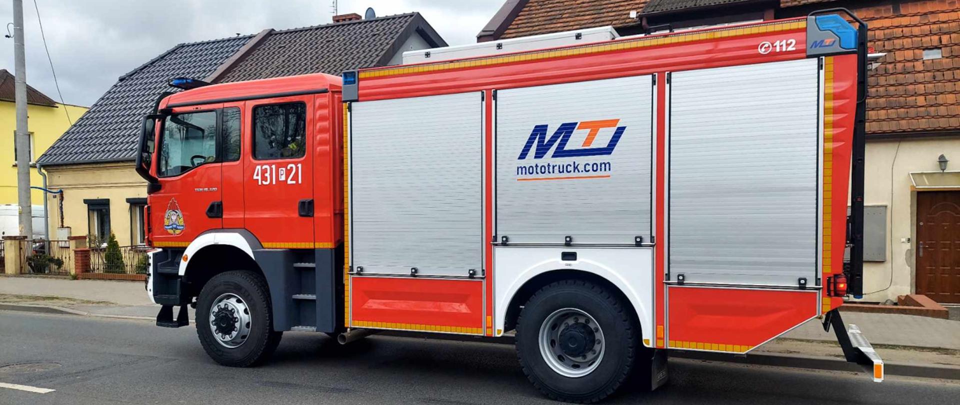 Wóz strażacki podczas zdarzenia. Samochód stoi na drodzy przy budynkach mieszkalnych.