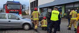 Parking przed sklepem. Na placu przed sklepem. Pomiędzy zaparkowanymi autami osobowymi strażacy udzielają kwalifikowanej pierwszej pomocy potrąconej kobiecie. Obok stoi policjant oraz dwóch mężczyzn w ubraniach cywilnych. W tle budynki, z lewej strony stoi wóz straży pożarnej. 