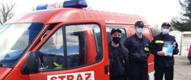 Dystrybucja ulotek. Trzech strażaków OSP na tle samochodu strażackiego. Strażacy trzymają w ręku ulotki.