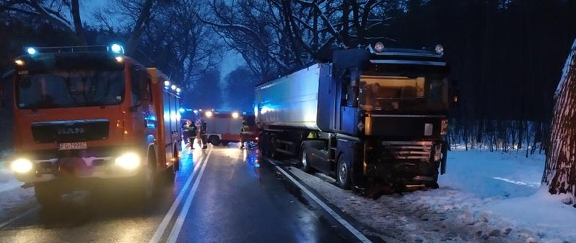 Zdjęcie przedstawia widok wzdłuż drogi krajowej numer 22. Panuje półmrok. Zdjęcie wykonane w osi drogi krajowej numer 22 w kierunku na Strzelce Krajeńskie. Po prawej stronie na poboczu zaraz przy pasie jezdni stoi samochód ciężarowy MAN, z naczepą do przewozu materiałów sypkich, kabina w kolorze czarnym, uszkodzona dolna część osłony silnika oraz częściowo reflektory. Na lewym pasie, patrząc od strony fotografującego, stoi średni samochód gaśniczy z włączonymi niebieskimi światłami ostrzegawczymi. Na dalszym planie, pomiędzy samochodami widać samochód ratownictwa technicznego oraz funkcjonariuszy Komendy Powiatowej PSP w Strzelcach Krajeńskich w ubraniach dowódczo-sztabowych. Panuje półmrok, na poboczach leży śnieg.