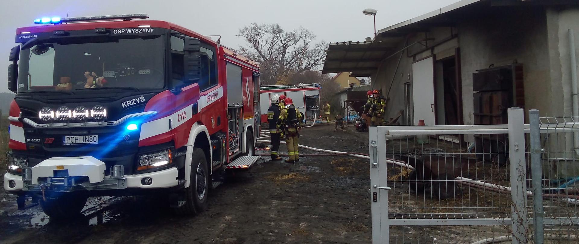 Zdjęcie przedstawia samochód strażacki oraz strażaków gaszących pożar w budynku inwentarskiego.
W tle budynek i inny samochód strażacki.
