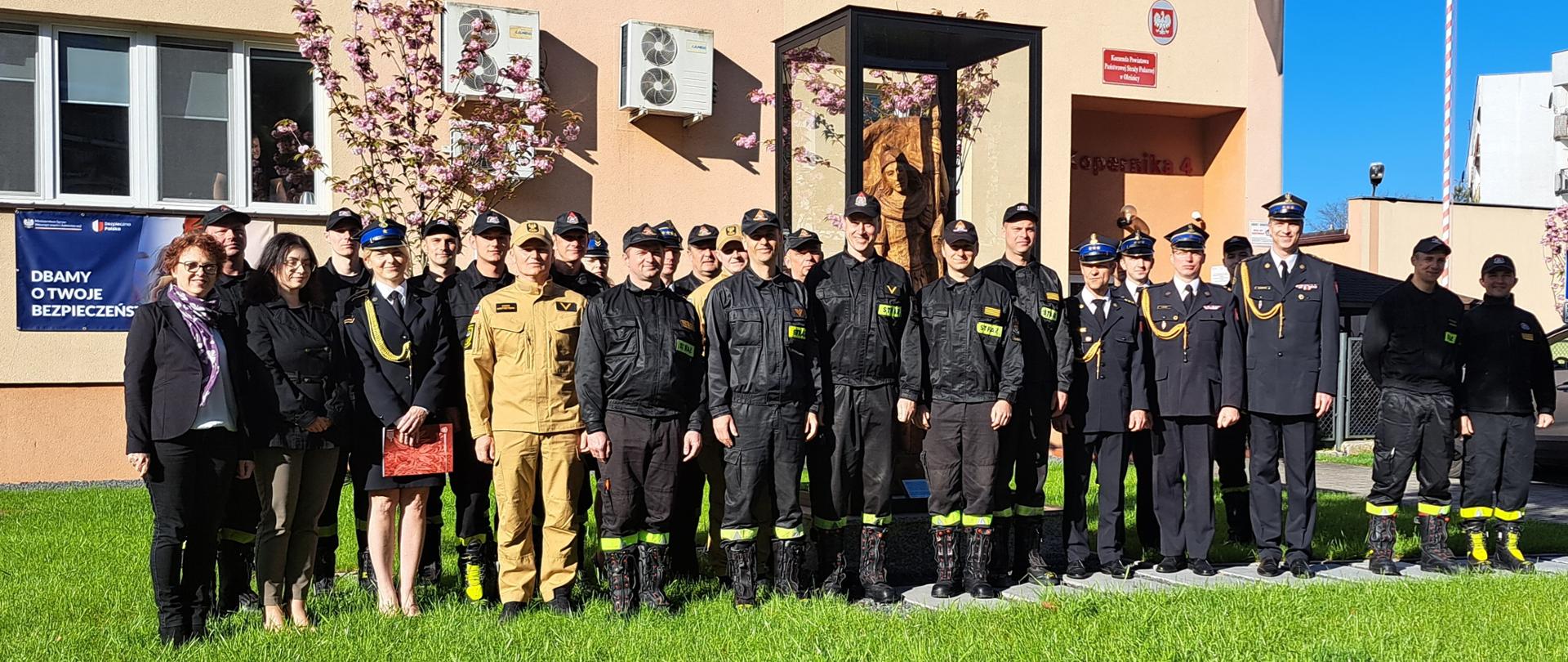 Zdjęcie grupowe oleśnickich strażaków przed budynkiem KP PSP Oleśnica. w tle figura Św. Floriana. 