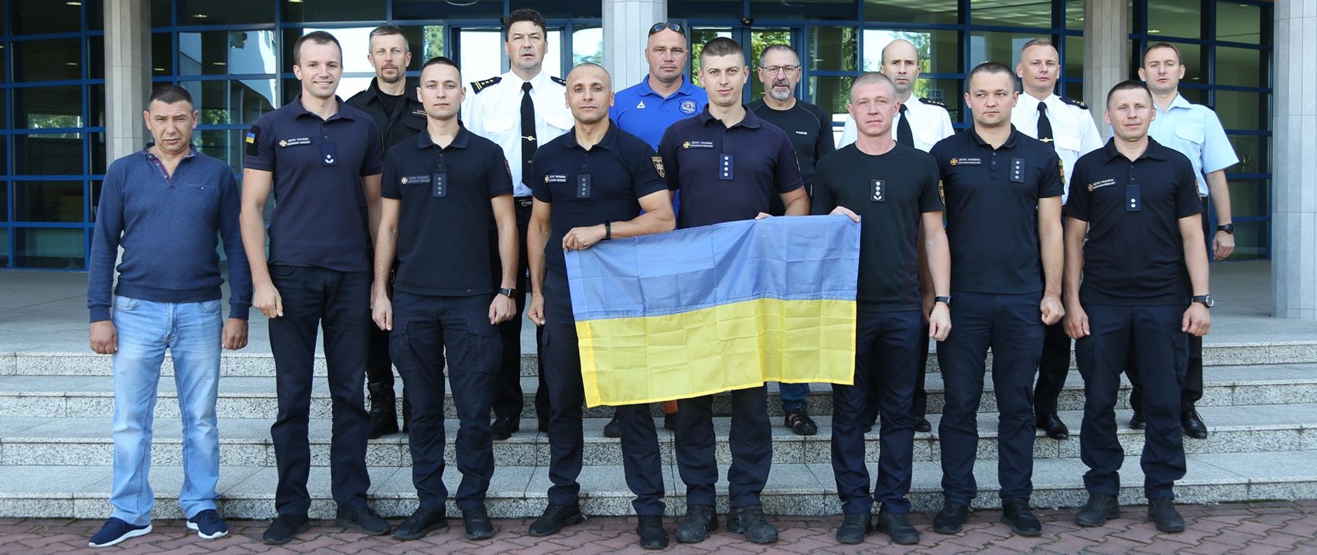 Przed budynkiem Szkoły polscy i ukraińscy strażacy pozują do grupowego zdjęcia. Trzech strażaków w pierwszym rzędzie trzyma flagę Ukrainy